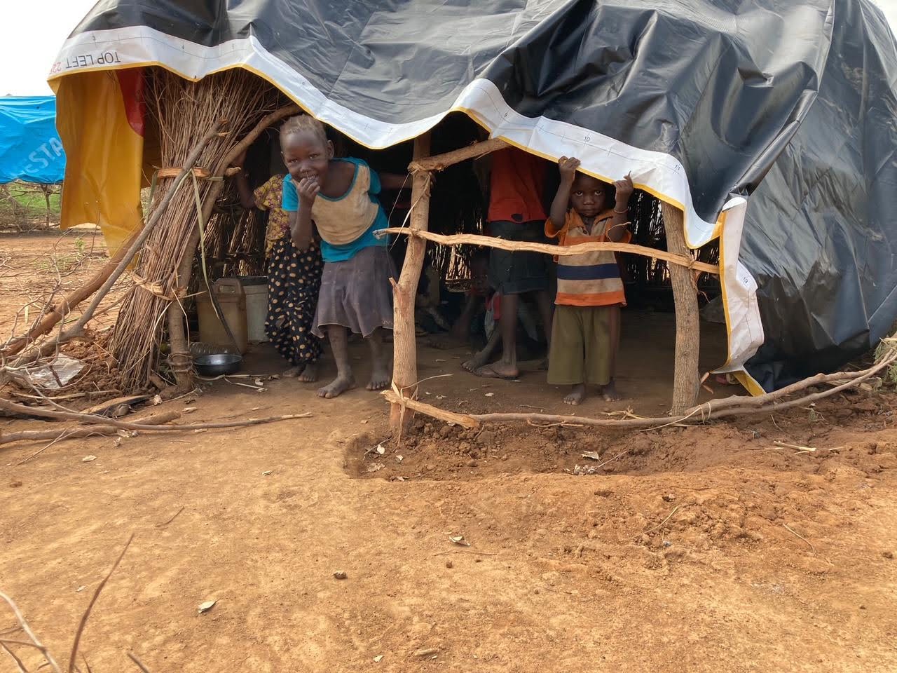 Sudanese children stand beneath Meadow Outdoor billboard tarp over their hut